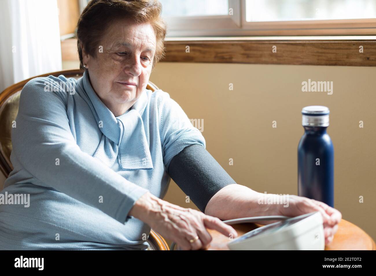 Kaukasische Frau über 70, die ihren Blutdruck zu Hause in der Nähe des Fensters überprüft.kurze Haare, blauer Pullover und kühle Wasserflasche. Stockfoto
