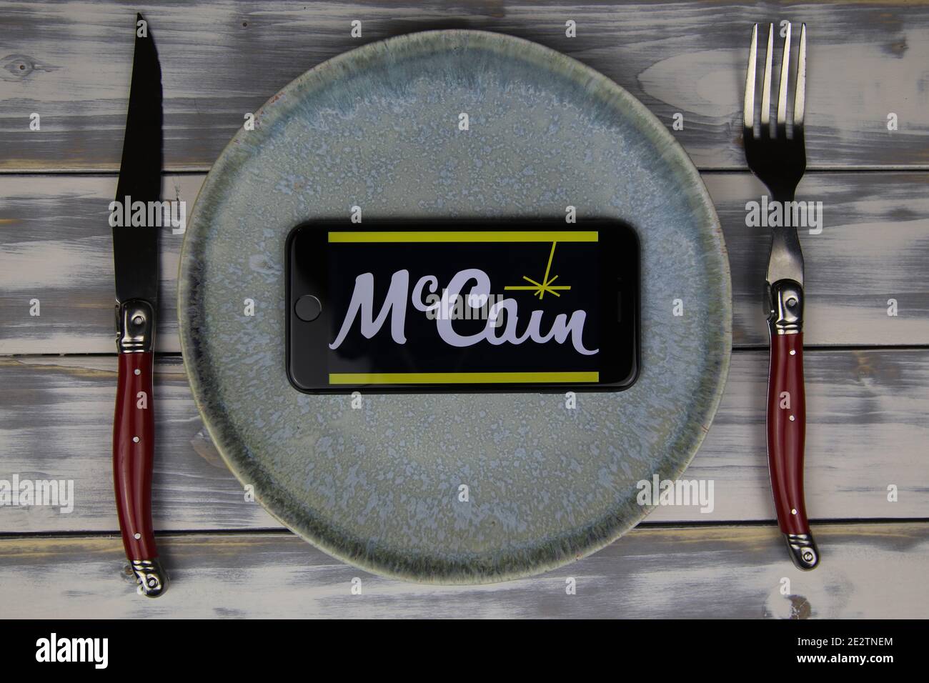 Viersen, Deutschland - 9. Mai. 2020: Nahaufnahme des Mobiltelefons auf Teller und Holztisch, Besteck mit Logo-Schriftzug der mccain Food Company (Schwerpunkt Mitte) Stockfoto