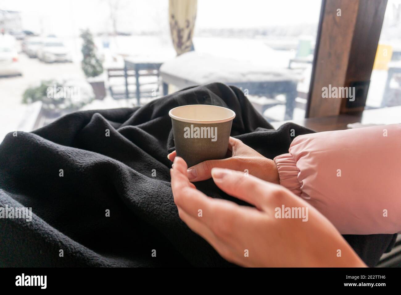 Eine Papiertasse Kaffee in den Händen eines Mädchens in einem Café, eine Theke mit Panoramafenster, ein Schild vor dem Fenster Stockfoto