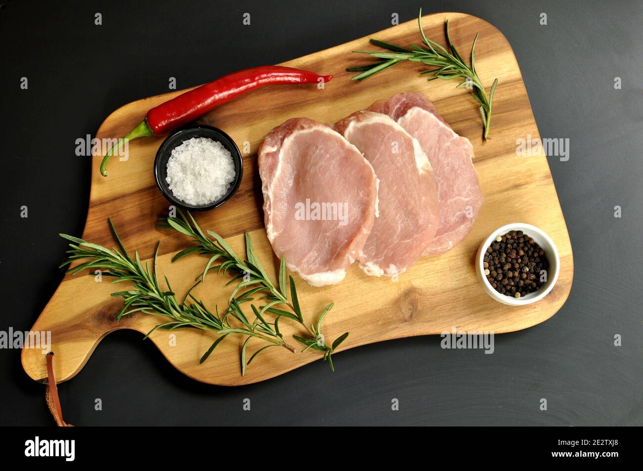 Roh frisch ungekocht geschnittenes Schweinefleisch Filetgericht mit Rosmarin, Pfeffer, Salz, rote Chili Pfeffer auf Holzbrett und schwarzem Hintergrund. Tisch auf der obersten Platte Stockfoto