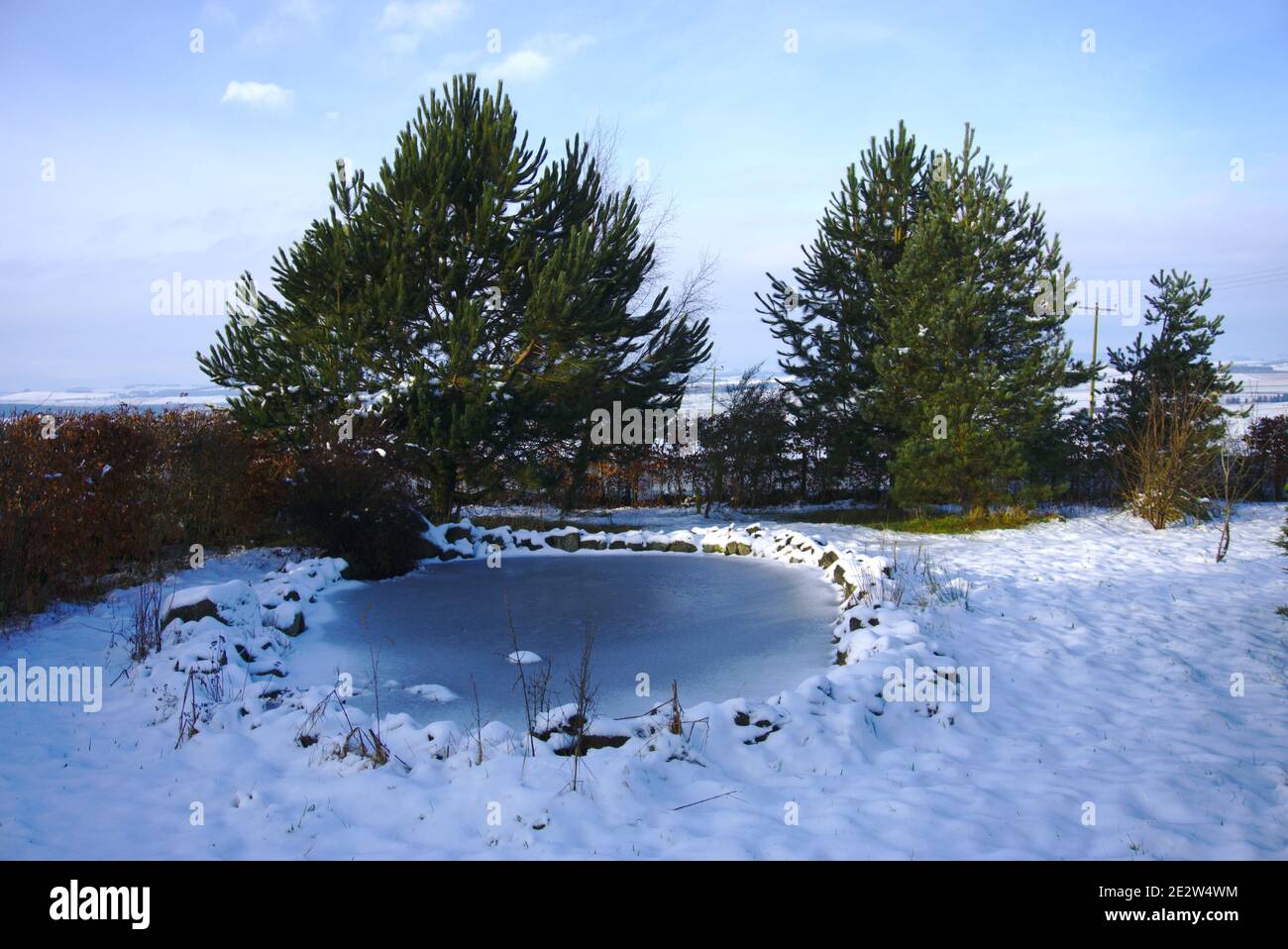 Gefrorener Teich und schneebedeckter Boden reflektieren blauen Himmel an einem klaren Wintertag in einem Garten in Berwickshire, Scottish Borders, Schottland, Großbritannien. Stockfoto