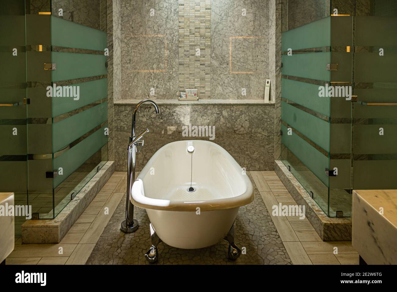 Eine freistehende Badewanne mit Klauenfüßen steht zwischen zwei mattierten Glasscheiben in einem marmorverkleideten Hotelbadezimmer in Grenada. Stockfoto