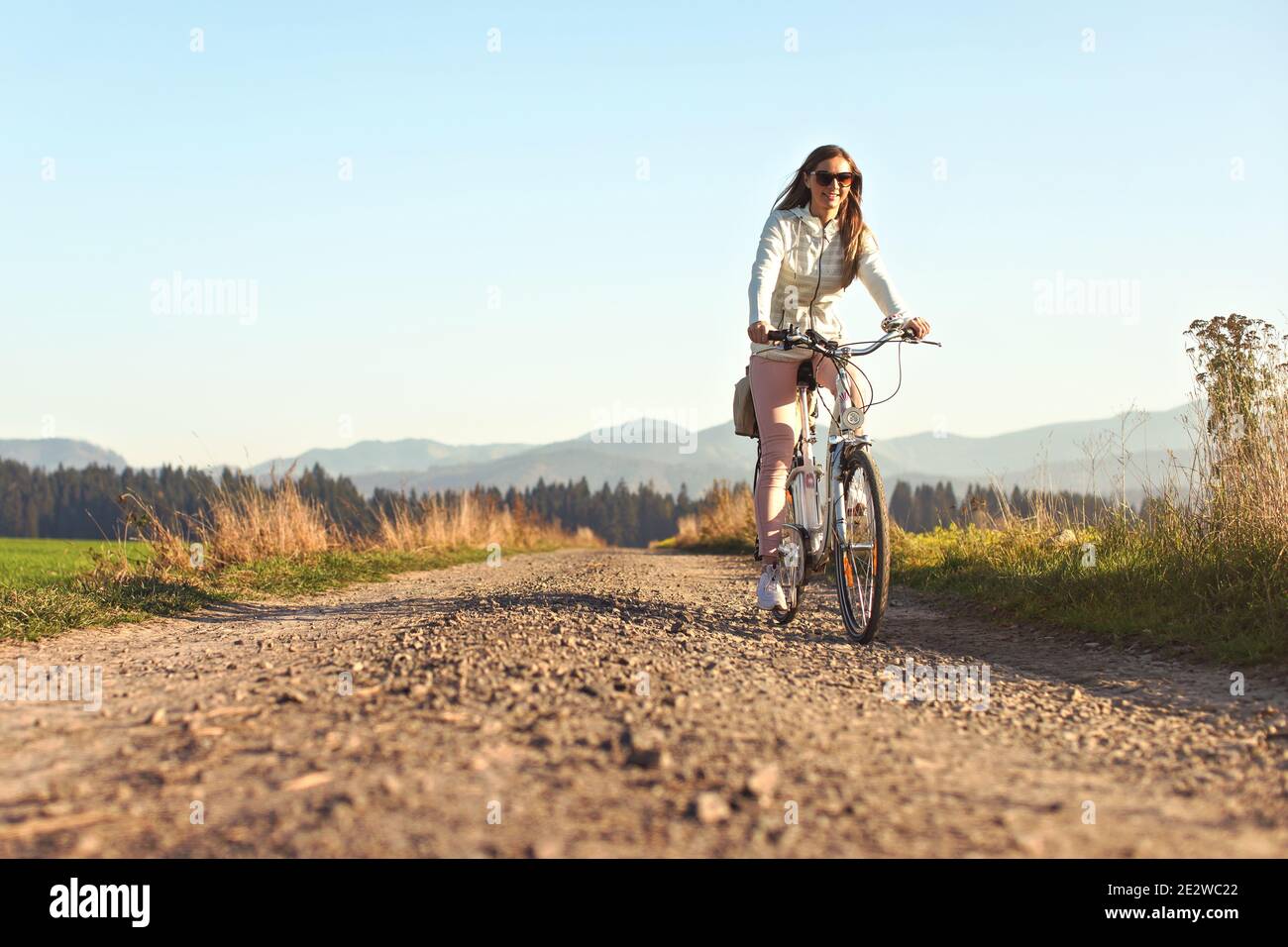 Junge Frau mit langen Haaren mit Sonnenbrille Fahrrad fahren auf staubigen Landstraße, Nachmittag Sonne beleuchtet Land Hintergrund Stockfoto