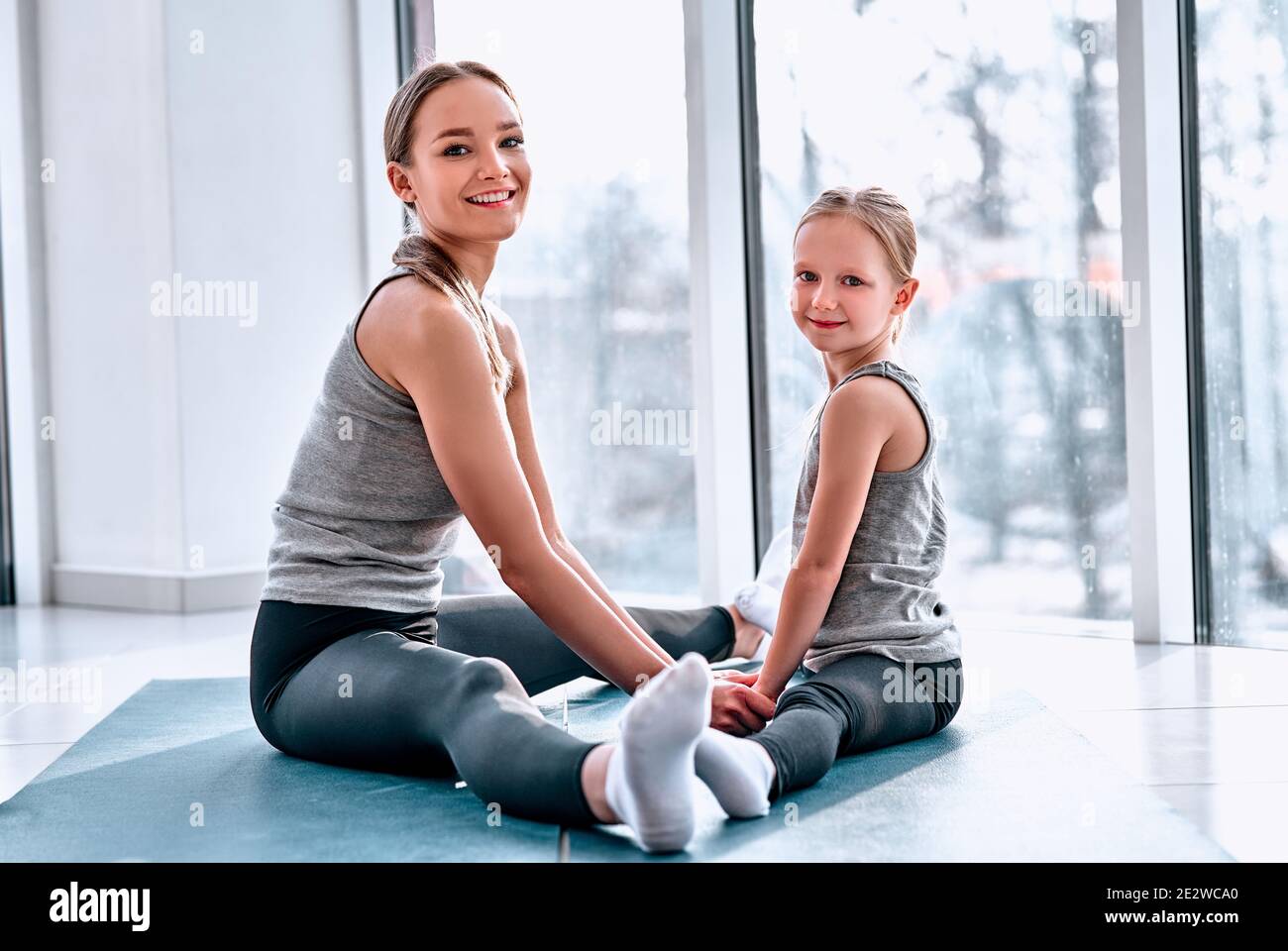 Mutter und Tochter machen Yoga. Familie in einem Fitnessraum. Kleines Mädchen mit Mutter in weißen T-Shirts und schwarzen Leggings. Mädchen sitzen auf einer grünen Matte Stockfoto
