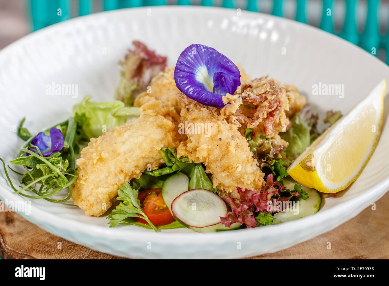 Gebutterter Fisch und grüner Salat mit Sauce und Zitrone, verziert mit Schmetterlingsblüten. Serviert auf einer weißen Keramikplatte. Stockfoto