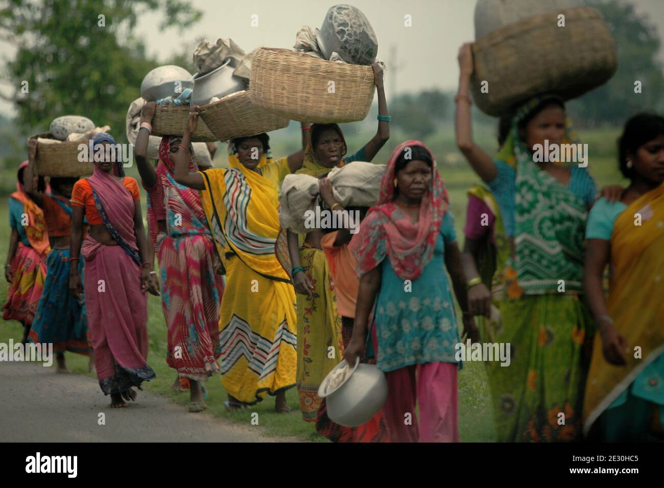 Frauen gehen in einer Schlange, tragen Körbe und Metallkrüge, während sie auf einer Straße in der ländlichen Gegend von Bihar, Indien, unterwegs sind. © Reynold Sumayku Stockfoto