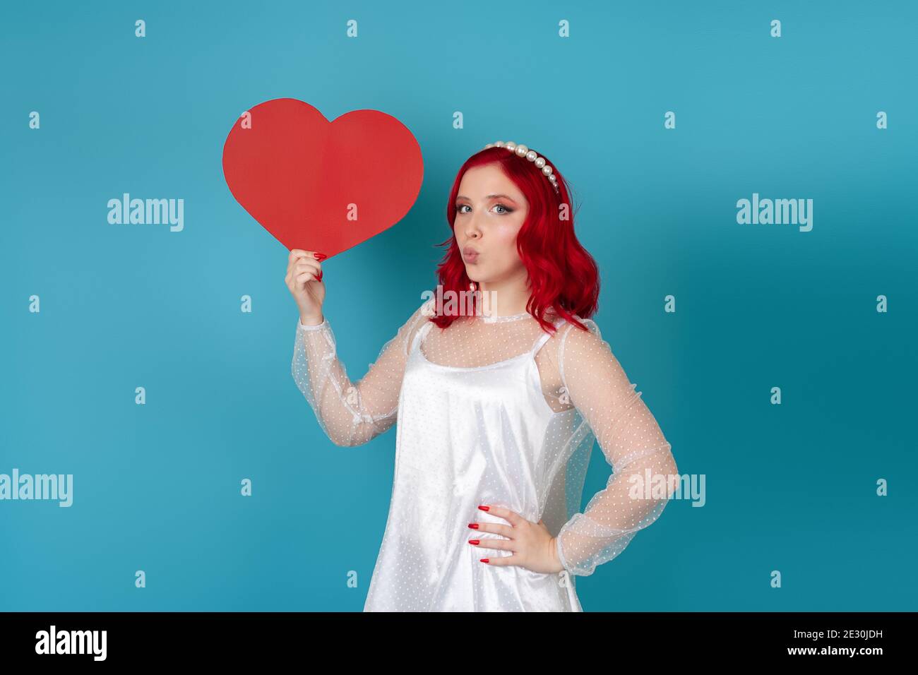 Charmante junge Frau in einem weißen Kleid und mit rot Haar hält ein großes rotes Papierherz und bläst ein Kiss isoliert auf einem blauen Hintergrund Stockfoto