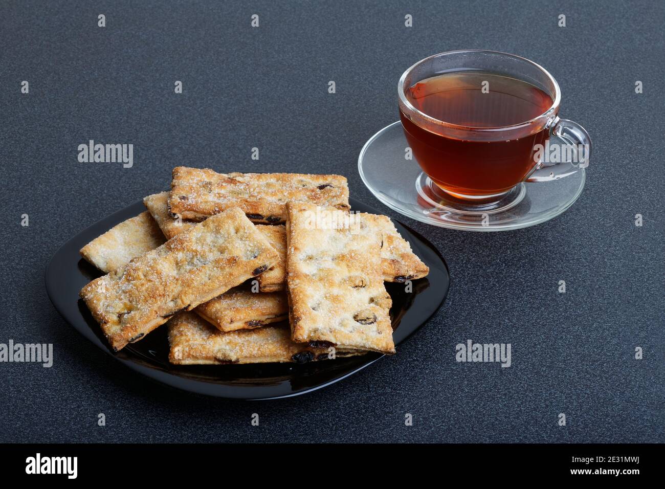 Blätterteig mit Rosinen auf einem schwarzen Teller und Tasse Von Tee auf einem dunklen Tisch Stockfoto