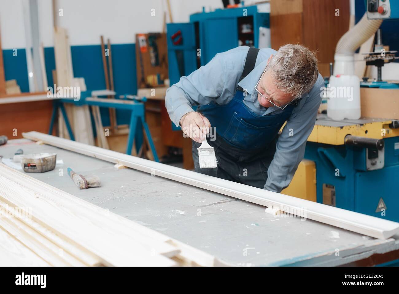Ein älterer Tischler in Overalls und Gläsern malt in einer Schreinerei mit einem Pinsel ein Holzbrett auf eine Werkbank. Stockfoto