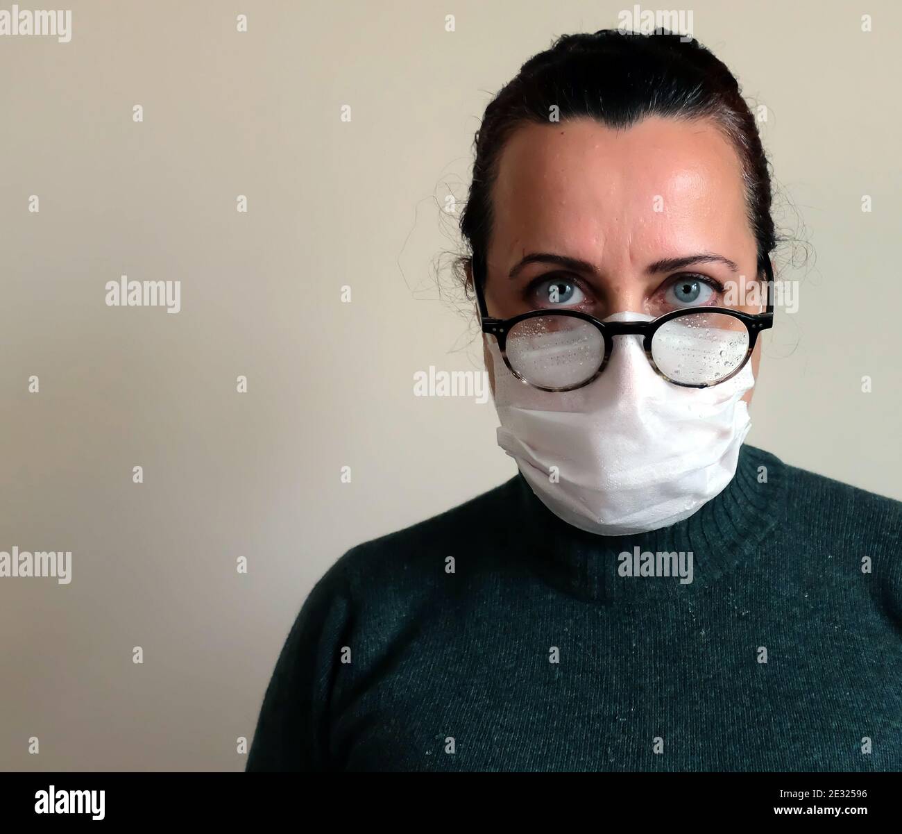 Frau in Brille mit weißer medizinischer Maske, Frau in weißer medizinischer Maske mit Brille mit Regentropfen, Frau in grünem Pullover vor Weiß stehend Stockfoto