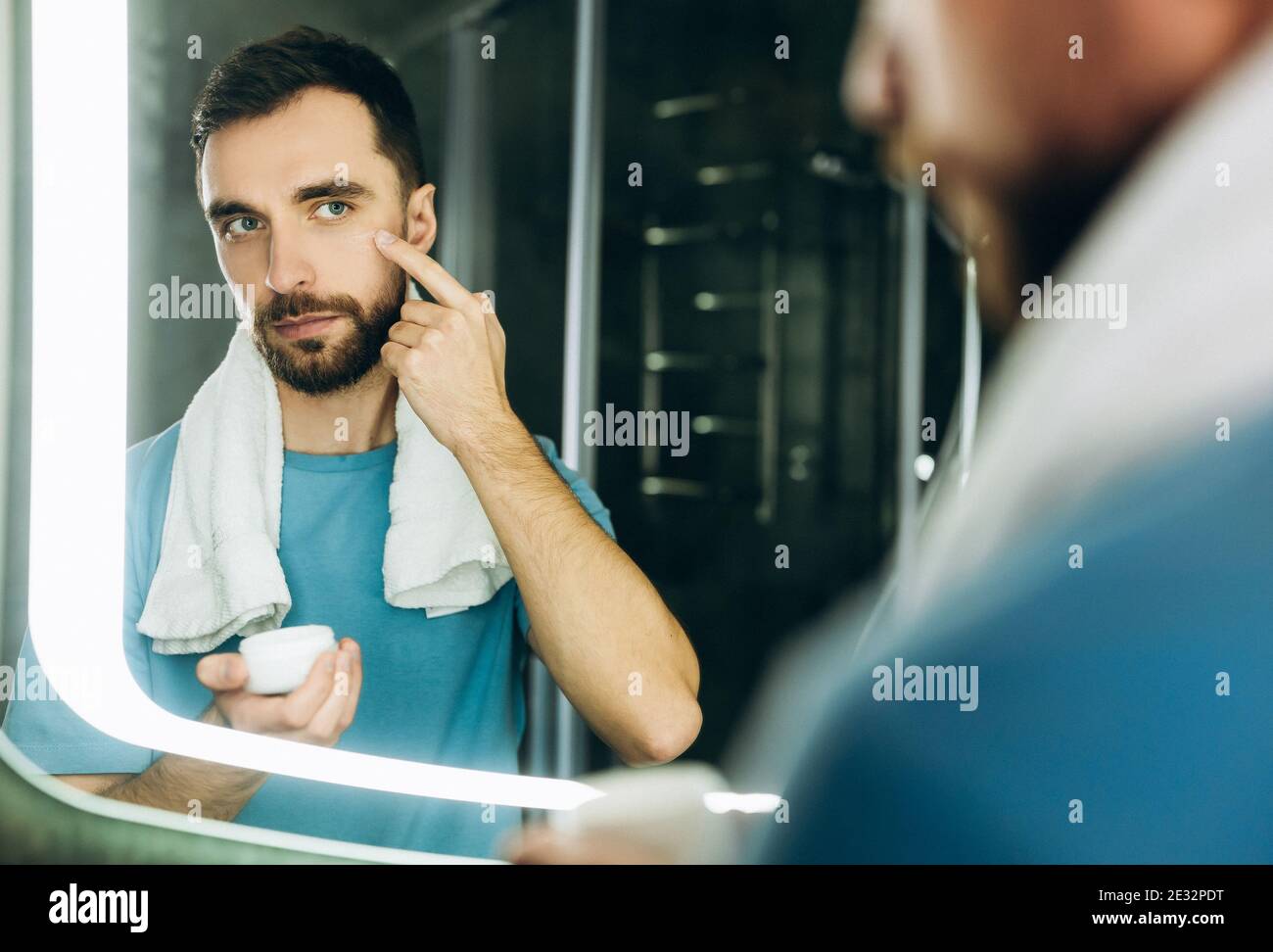 Nahaufnahme eines hübschen jungen Mannes mit einem Handtuch im Nacken, das vor dem Spiegel steht und feuchtigkeitsspendende Creme auf sein Gesicht auftragen. Stockfoto