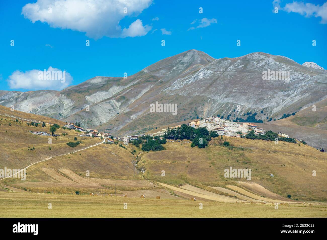 Die Stadt Castelluccio di Norcia von der Hochebene Pian Grande aus gesehen. Sommersaison, gelbes Gras. Castelluccio wurde vor kurzem von einem Erdbeben getroffen Stockfoto