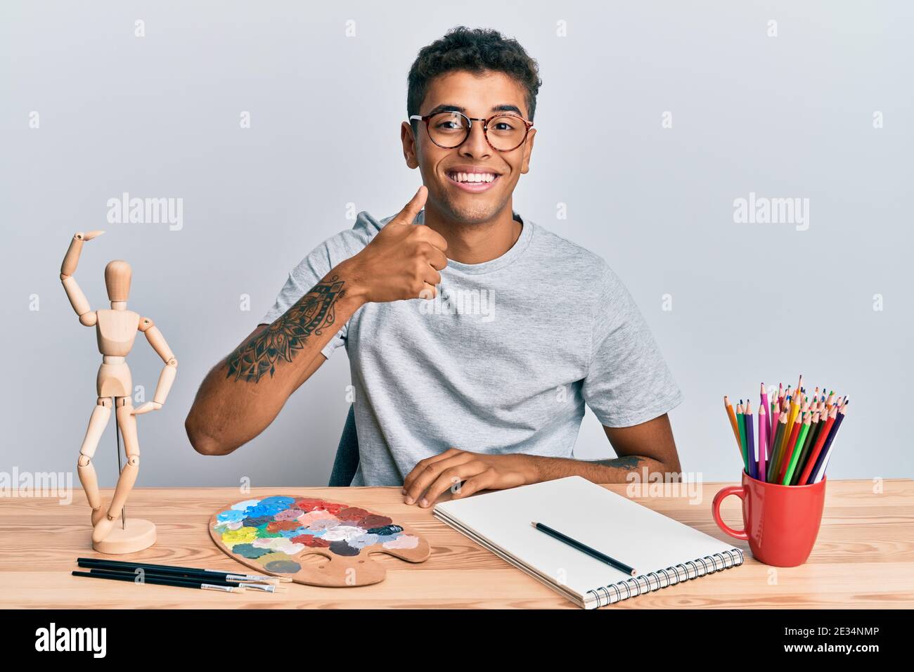 Junge gut aussehende afroamerikanische Mann Maler sitzende Palette und Kunst-Übungspuppe tun glücklich Daumen nach oben Geste mit der Hand. Genehmigung Ausdruck Blick auf t Stockfoto