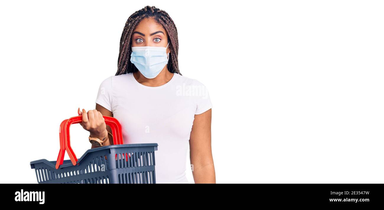 Junge afroamerikanische Frau mit Zöpfen tragen Warenkorb und medizinische Maske erschrocken und erstaunt mit offenem Mund für Überraschung, Unglauben Gesicht Stockfoto