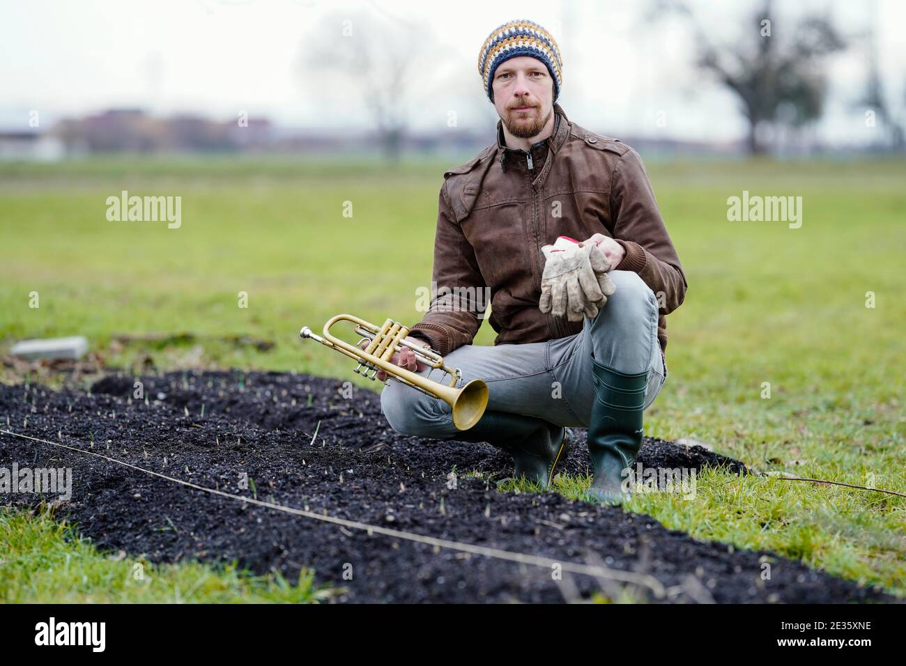 Mannheim, Deutschland. Januar 2021. Johannes Stange, ein Jazztrompeter und angehende Bio-Bauer, sitzt mit Trompete und Arbeitshandschuhen an einem Knoblauchpflanzenbett. Mit Blick auf die Absage von Konzerten aufgrund der Corona ändert er seine Karriere und schafft eine zweite wirtschaftliche Stütze mit Gartenarbeit. (An dpa-KORR: 'Keine Konzerte: Musiker schaffen zweite Stütze bei Gärtnern') Quelle: Uwe Anspach/dpa/Alamy Live News Stockfoto