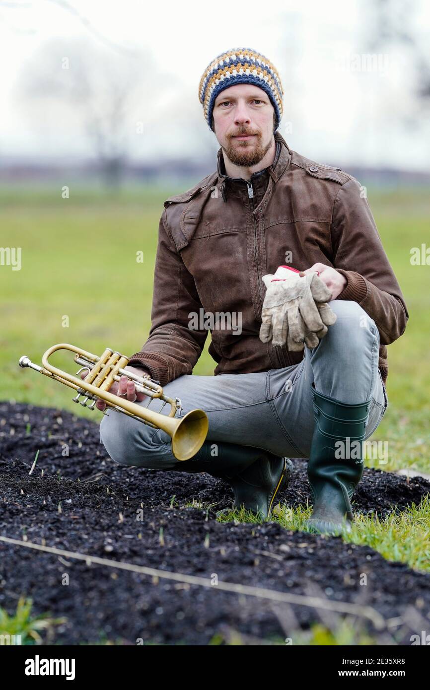 Mannheim, Deutschland. Januar 2021. Johannes Stange, ein Jazztrompeter und angehende Bio-Bauer, sitzt mit Trompete und Arbeitshandschuhen an einem Knoblauchpflanzenbett. Mit Blick auf die Absage von Konzerten aufgrund der Corona ändert er seine Karriere und schafft eine zweite wirtschaftliche Stütze mit Gartenarbeit. (An dpa-KORR: 'Keine Konzerte: Musiker schaffen zweite Stütze bei Gärtnern') Quelle: Uwe Anspach/dpa/Alamy Live News Stockfoto
