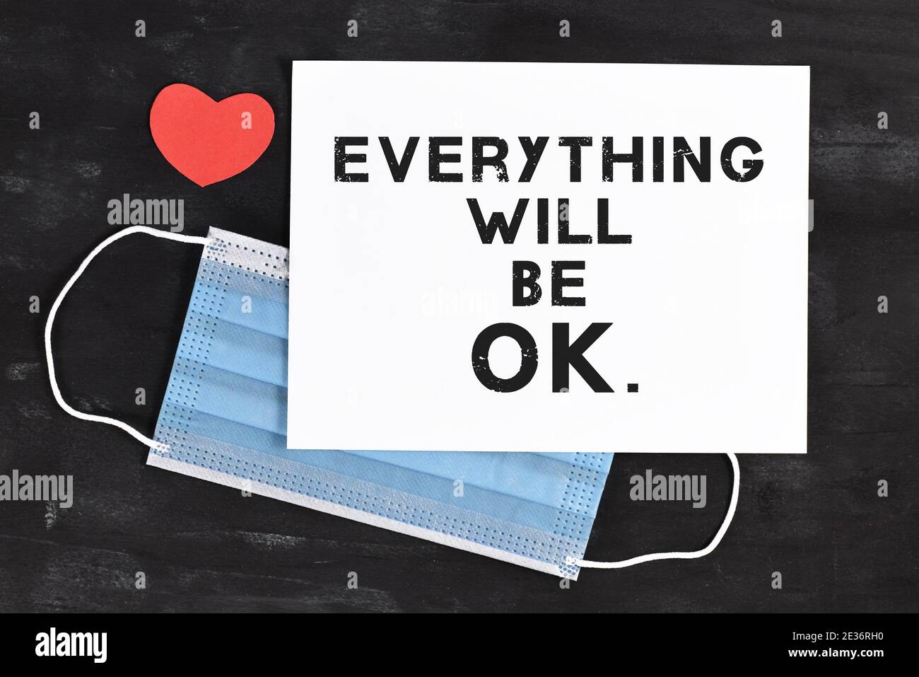 Text „alles wird in Ordnung sein“ auf weißem Papier davor Der schützenden Gesichtsmaske mit rotem Herz auf dunklem Hintergrund Stockfoto