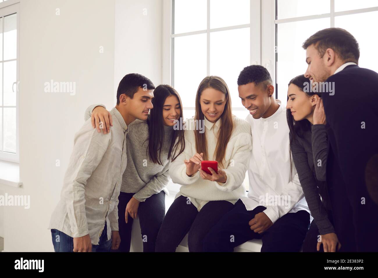 Multiethnische Gruppe von Menschen, die auf der Fensterbank sitzen und auf den Bildschirm eines Mobiltelefons schauen. Stockfoto