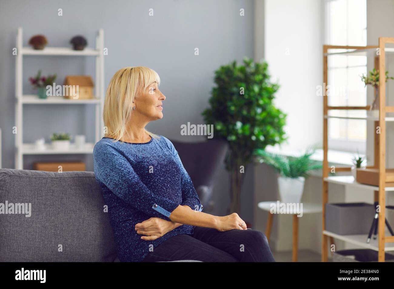 Nachdenklich lächelnde ältere Frau, die aus dem Fenster blickt und auf dem Sofa im gemütlichen Wohnzimmer sitzt. Stockfoto