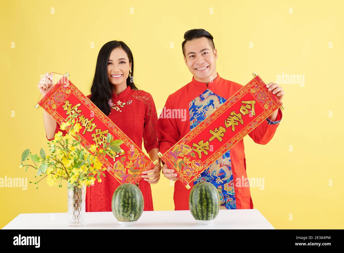Glücklich junge asiatische Freund und Freundin in traditionellen Kleidern zeigen Wanddekorationen für Lunar New Year mit der Aufschrift „Best Wishes“ Stehen bei Stockfoto