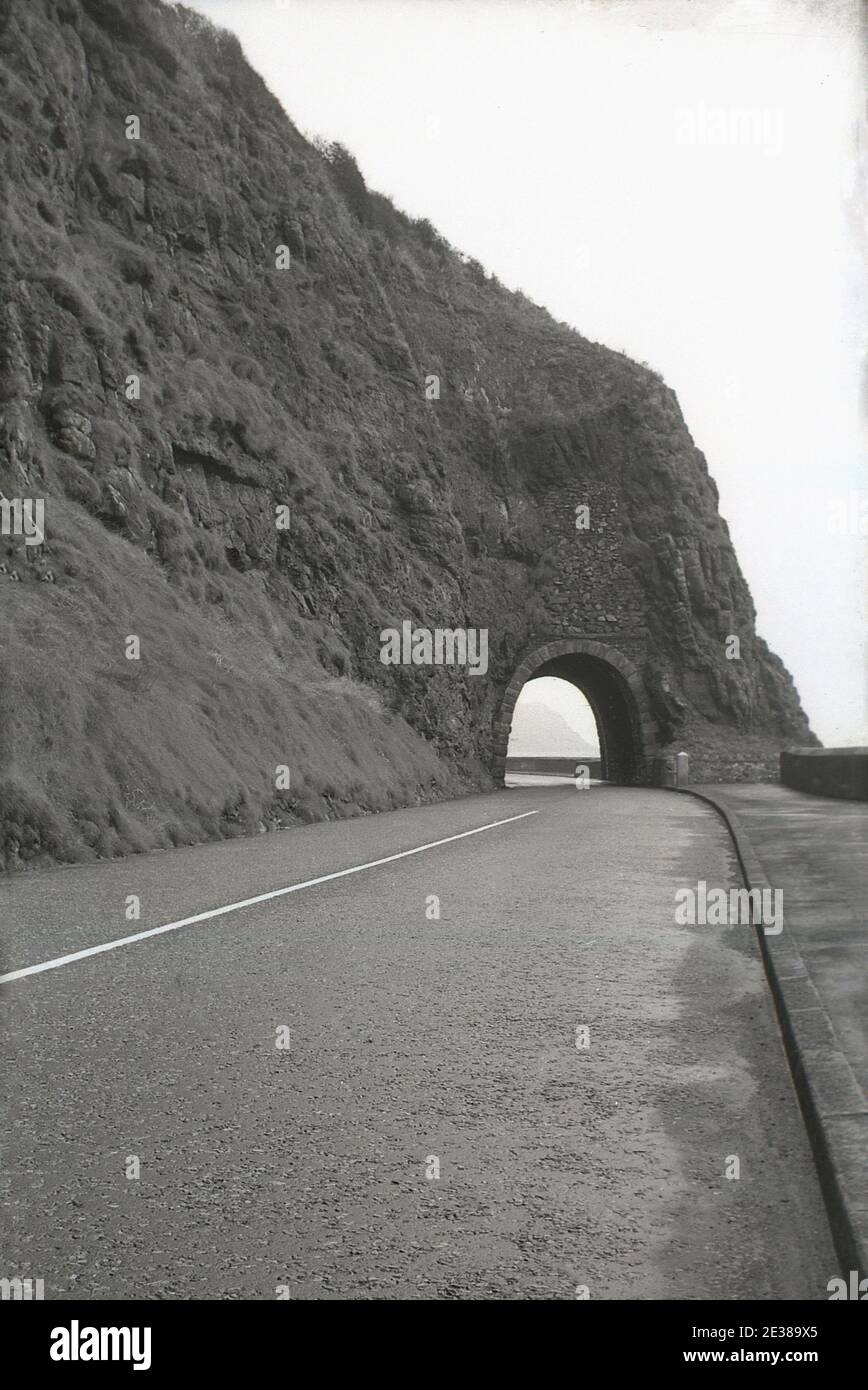1950er Jahre, historisch, in Larne, am Anfang der Causeway Küstenstraße (A2), geht die Straße unter dem Blackcave Tunnel oder "Black Arch", eine enge Öffnung oder Tunnel aus der überhängenden Klippe geschnitten, Larne, Antrim, Nordirland. Es war die Idee des Bauingenieurs William bald, der den Spielraum für den Bau einer Straße entlang der Kante der Klippe sah, die zwischen 1832 und 1842 stattfand und eine große Leistung für seine Zeit war. Die Straße blieb bis in die späten 1960er Jahre weitgehend unverändert. Stockfoto