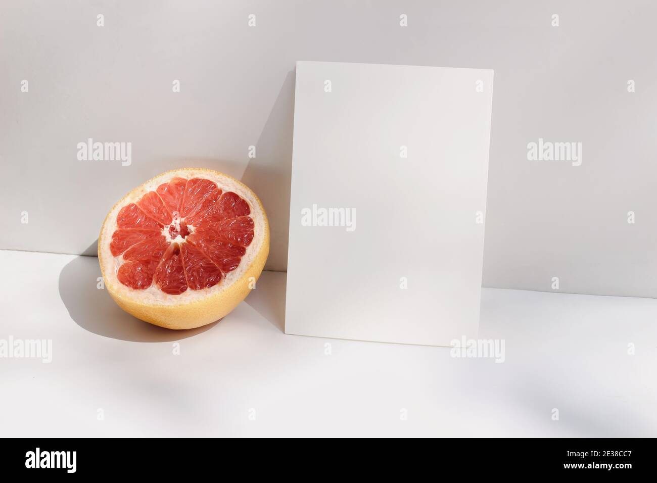 Moderne Sommer Schreibwaren Stillleben Szene. Nahaufnahme von geschnittenen Grapefruitfrüchten auf weißem Tischhintergrund im Sonnenlicht. Leere Papierkarte, Einladung Mockup Stockfoto