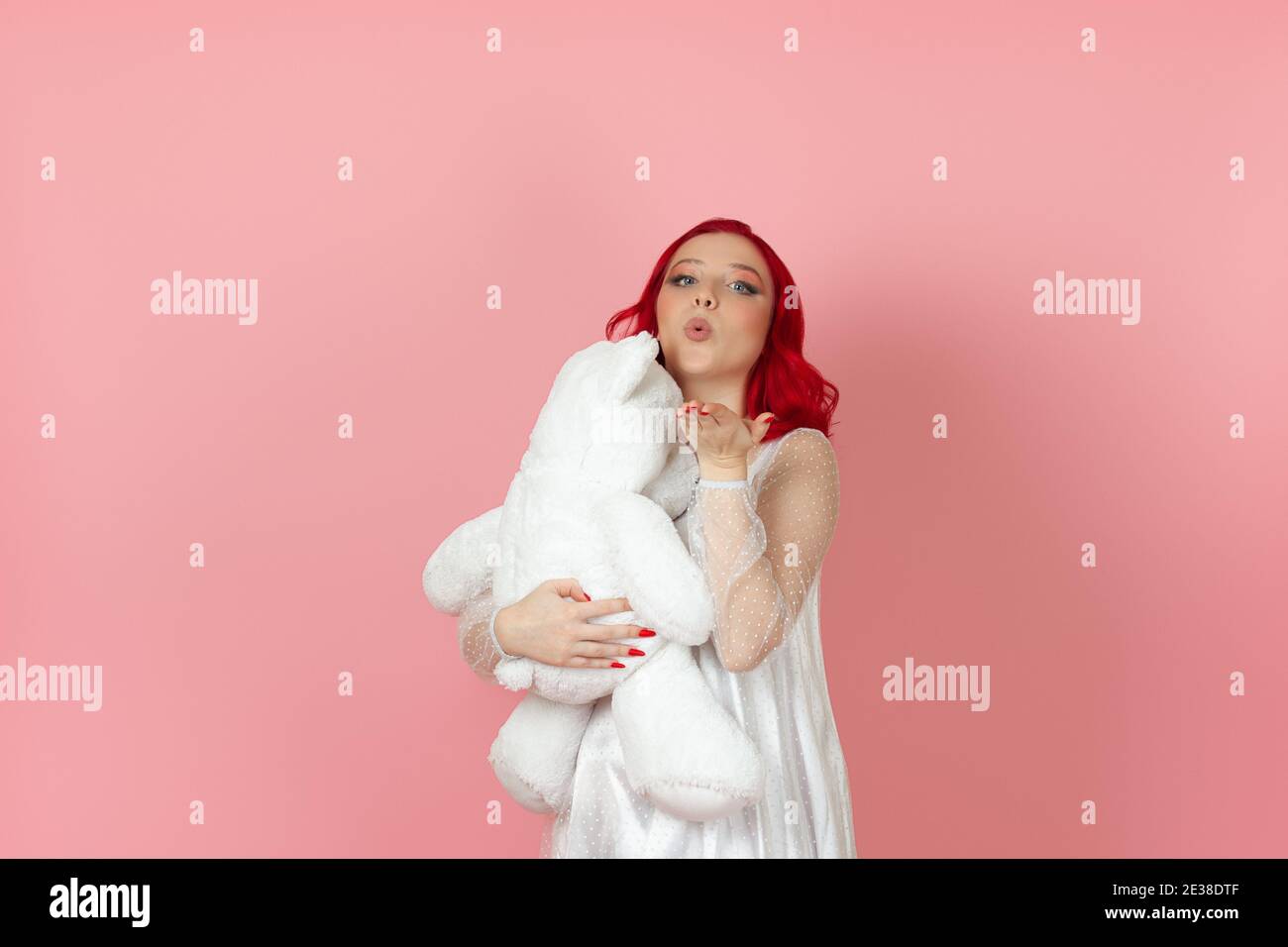 Nahaufnahme eine niedliche junge Frau in einem weißen Kleid und mit roten Haaren bläst einen Kuss und umarmt großen weißen Teddybär isoliert auf einem rosa Hintergrund. Stockfoto