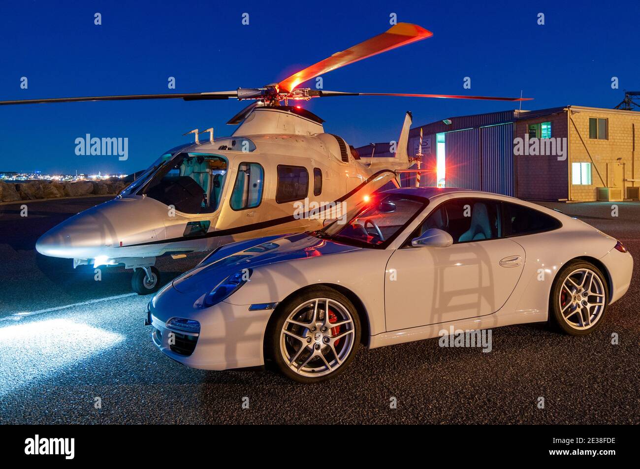 Ein Agusta 109 Power Executive Hubschrauber bei Dämmerung neben einem Porsche 911 Auto. Stockfoto