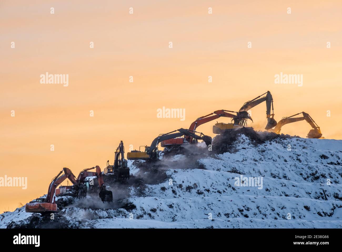 Silhouetten von vielen Baggern, die auf einem riesigen Berg arbeiten Eine Müllhalde vor dem Hintergrund eines orangefarbenen Sonnenaufgangs Oder Sonnenuntergang Himmel im Winter Stockfoto