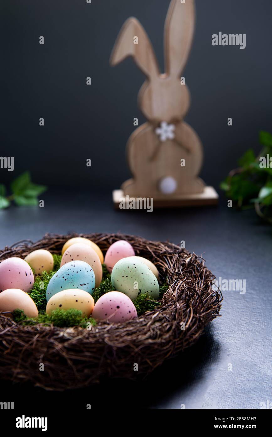 Ein Weidennest mit Moos und farbigen Ostereiern und Holzhasen-Kaninchen und grüne Pflanze hinterlassen auf dunklem Hintergrund. Frohe Ostern. Dunkle Stimmung Stockfoto