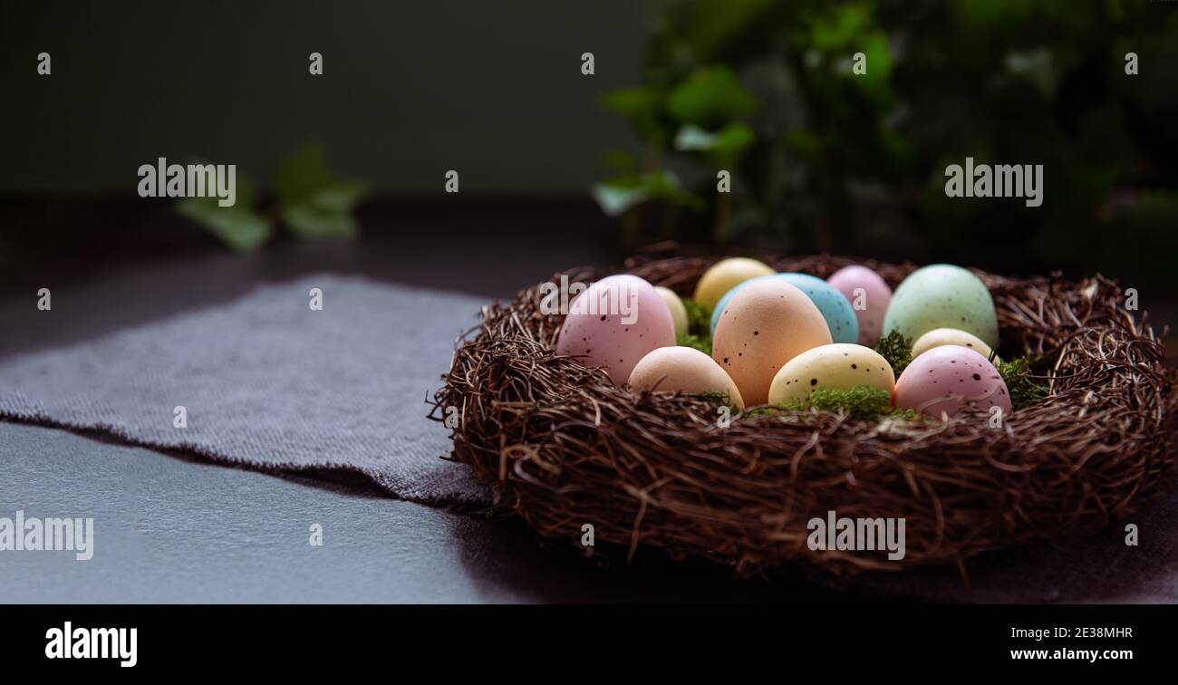 Ein Weidennest mit Moos und farbigen Ostereiern auf grauer Leinenserviette und wenigen grünen Blättern auf dunklem Grund. Dunkle Stimmung. Frohe Ostern Stockfoto