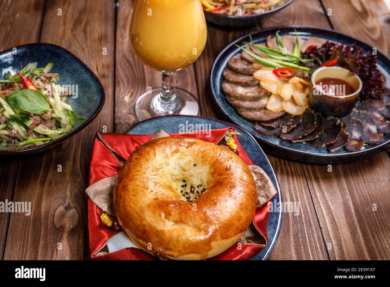 Serviert werden verschiedene Gerichte mit Gemüsescheiben, Fleischstücken, Salaten, Orangensaft und einem Brötchen auf braunem Holzhintergrund. Stockfoto