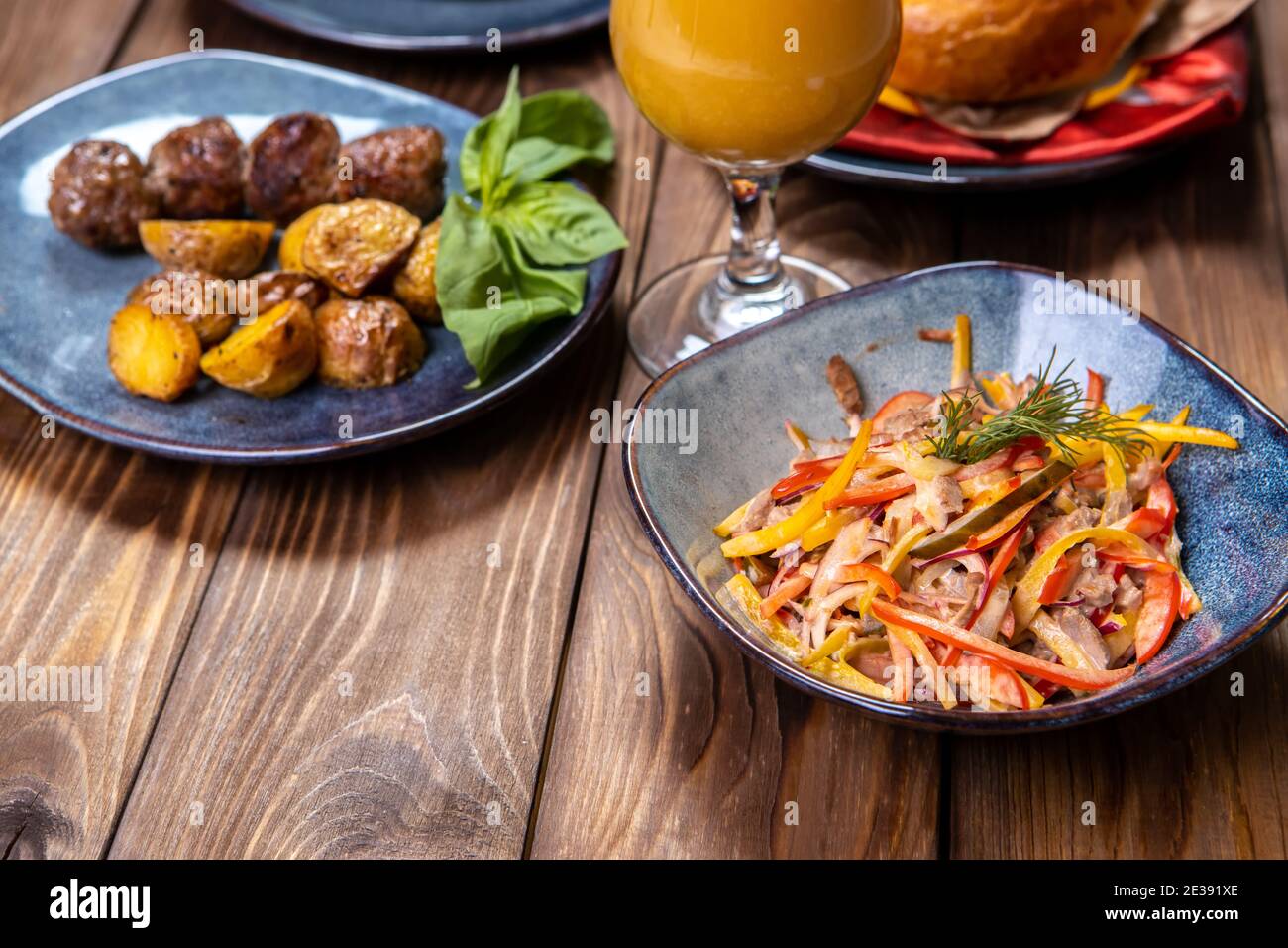 Eine Tasse mit einem Salat aus Gemüse gewürzt mit Sauce, frischem Gebäck, Orangensaft und Bratkartoffeln mit Koteletts auf braunem Holzhintergrund. Stockfoto