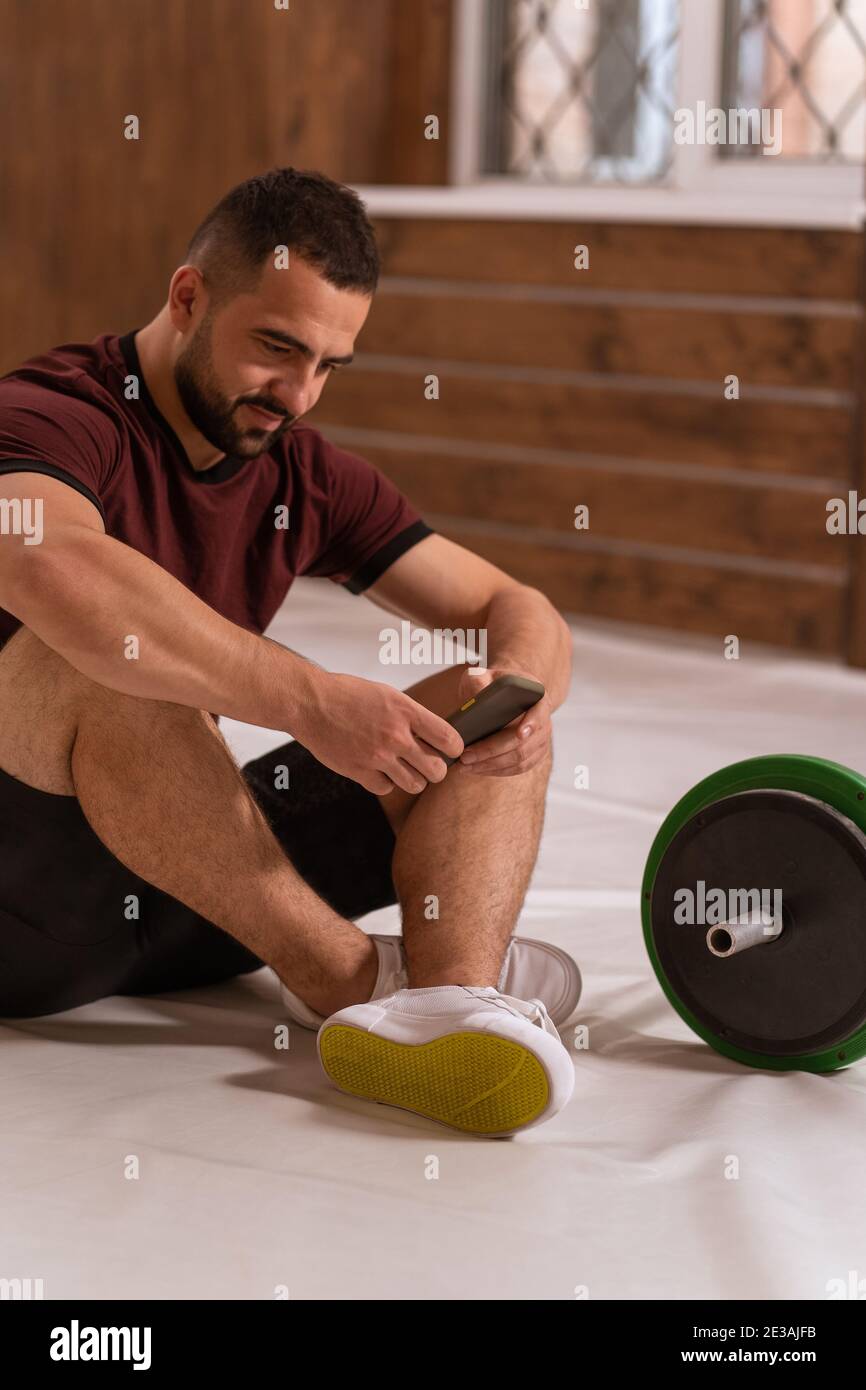 Schöner Mann sitzt auf einem Boden mit Smartphone in den Händen und schwarz-grün-Fitness-Langhantel, Geräte für Krafttraining Konzept. Sport Stockfoto