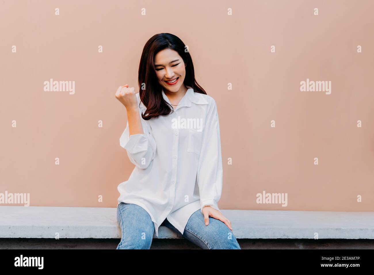 Jubelnde junge asiatische Mädchen hält ihre Fäuste fest wie gewinnen großen Preis, sitzt auf Betonbank mit Wüste Sand farbigen Wand im Hintergrund tragen lässige weiße Hemd und Jeans Kleidung Stockfoto
