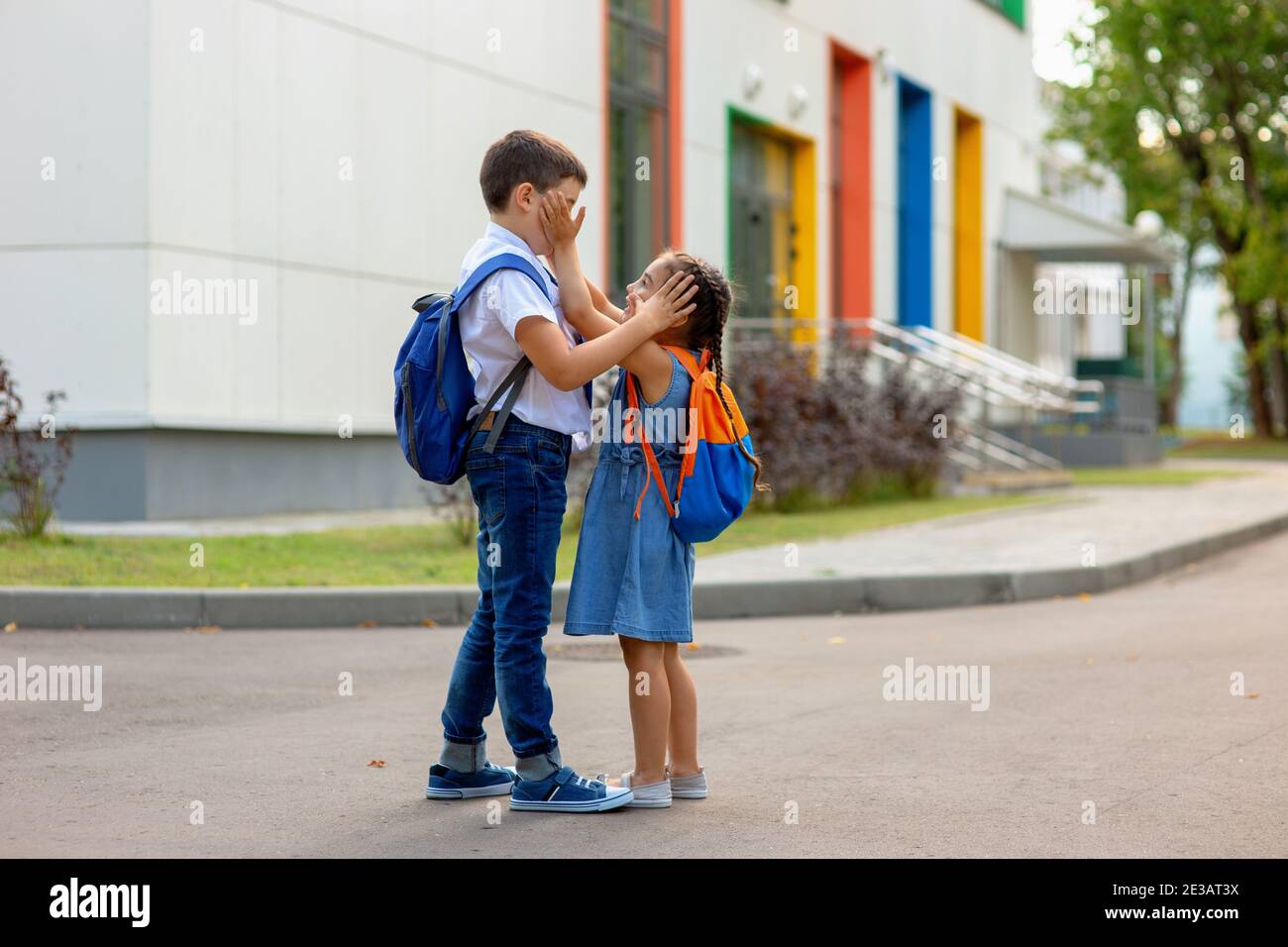 Ein kleines Mädchen und ein Bruder mit Rucksäcken halten jeden fest Andere an den Handflächen ihrer Gesichter in der Nähe der Schule Gebäude Stockfoto