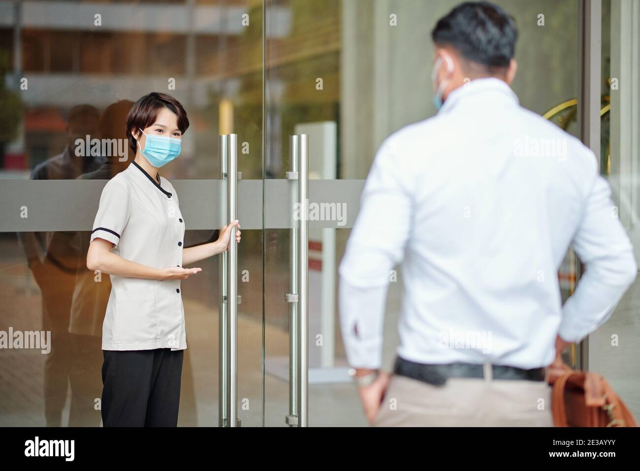 Junge weibliche Hotel Concierge in medizinische Maske Tür öffnen für Gast und ihn innen einladen Stockfoto