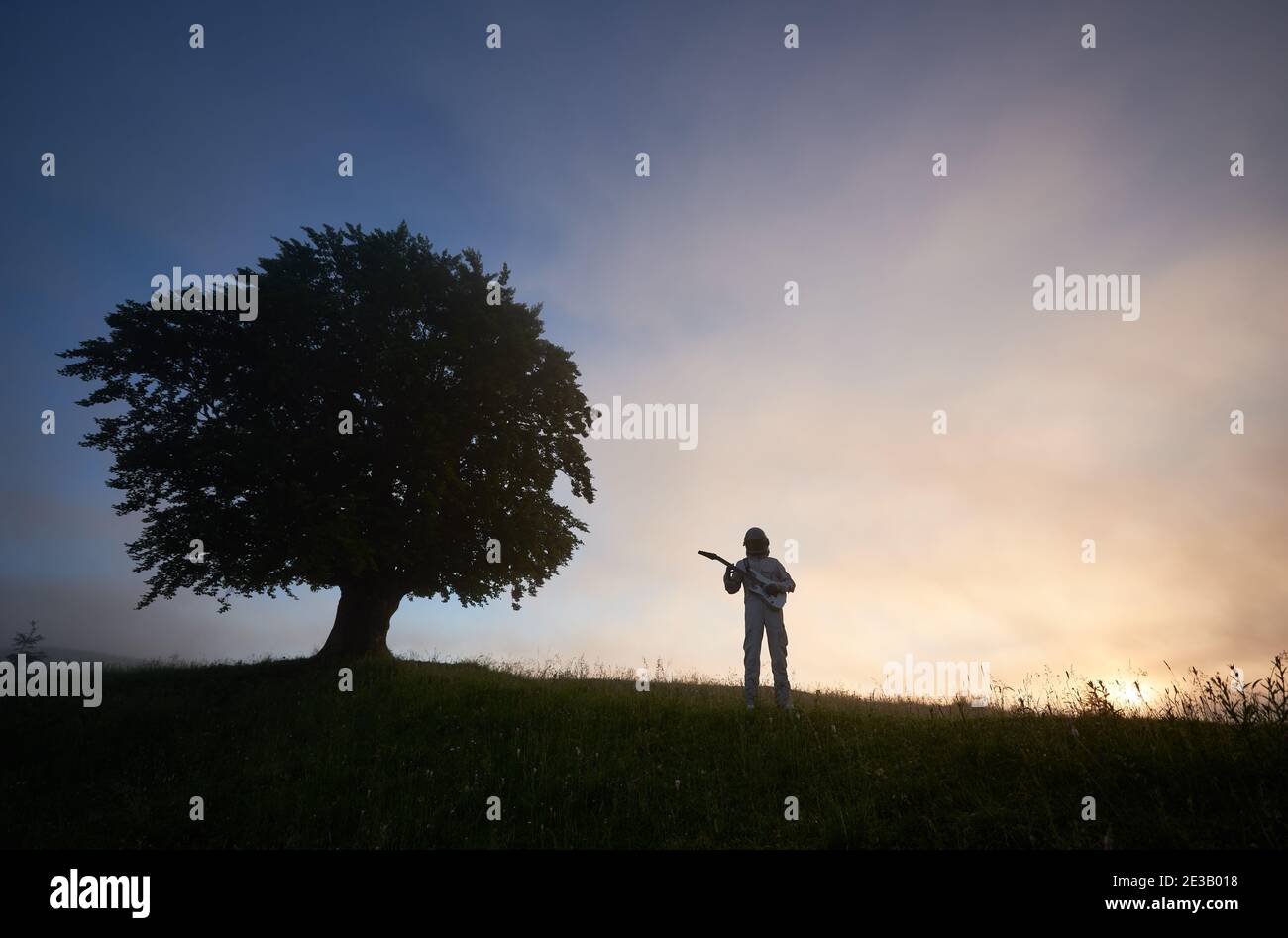 Horizontale Momentaufnahme von zwei Silhouetten auf violettem Himmel Hintergrund, von einem Raumfahrer im Raumanzug, der Gitarre spielt und einem großen alten Baum auf der rechten Seite. Konzept von Musik, Kosmonauten und Natur. Stockfoto
