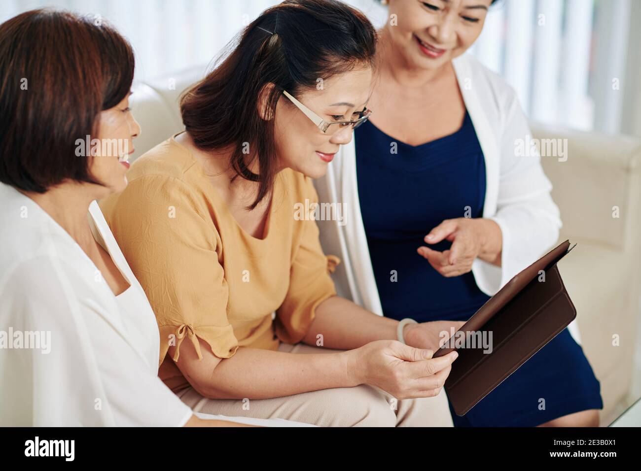 Gruppe von lächelnden älteren vietnamesischen Frauen diskutieren Fotos auf Tablet Computer, wenn man zu Hause auf dem Sofa sitzt Stockfoto