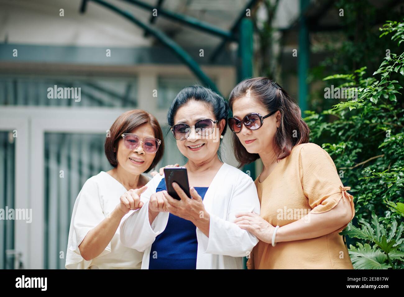 Fröhliche ältere asiatische Frauen in Sonnenbrillen stehen im Hinterhof und Überprüfen von „Gefällt mir“-Angaben auf Fotos, die sie in sozialen Medien hochgeladen haben Stockfoto
