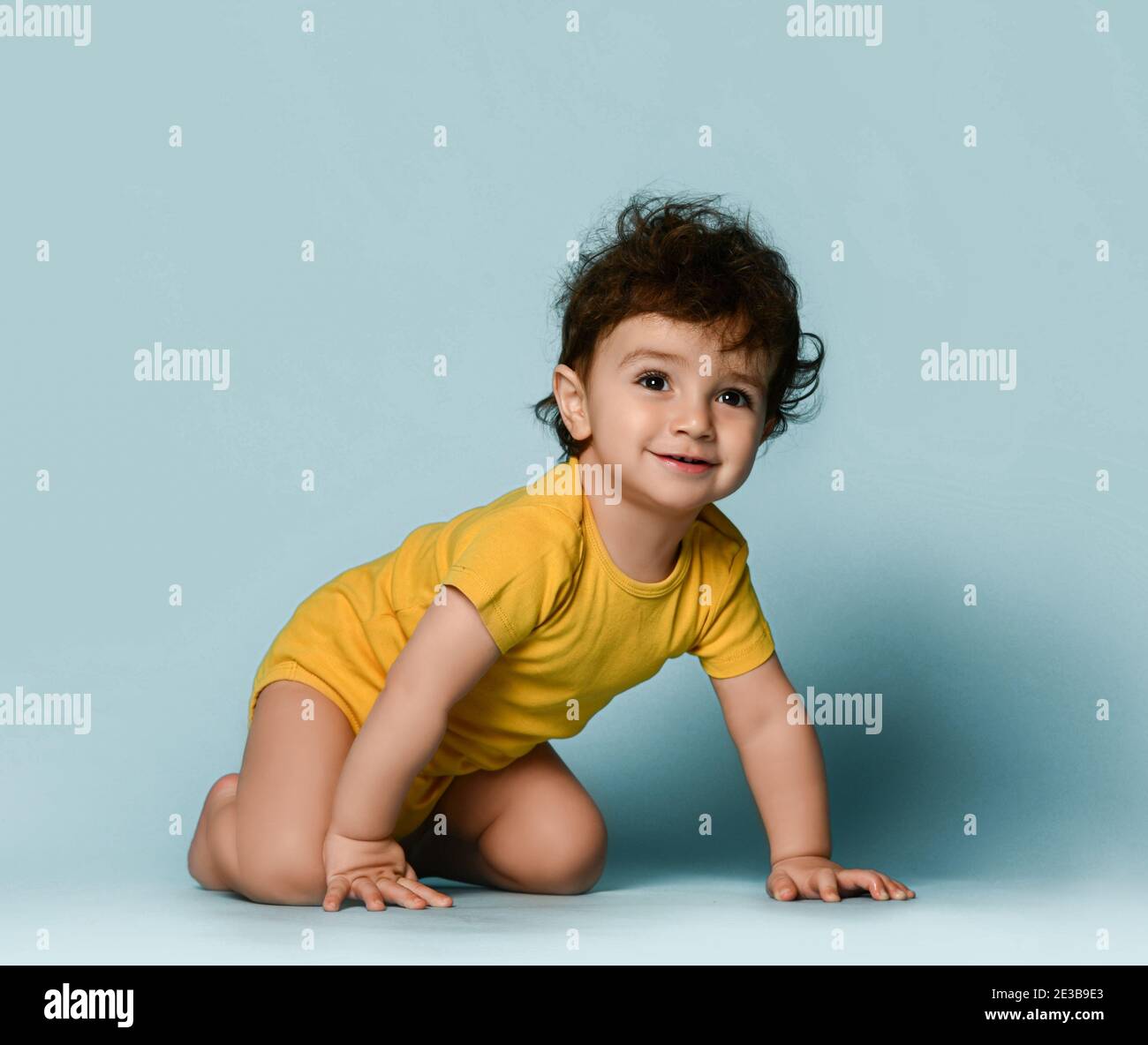 Kleine nette positive lockige Baby junge Kleinkind in gelb bequem Jumpsuit kriecht auf dem Boden und schaut auf die Kamera Stockfoto