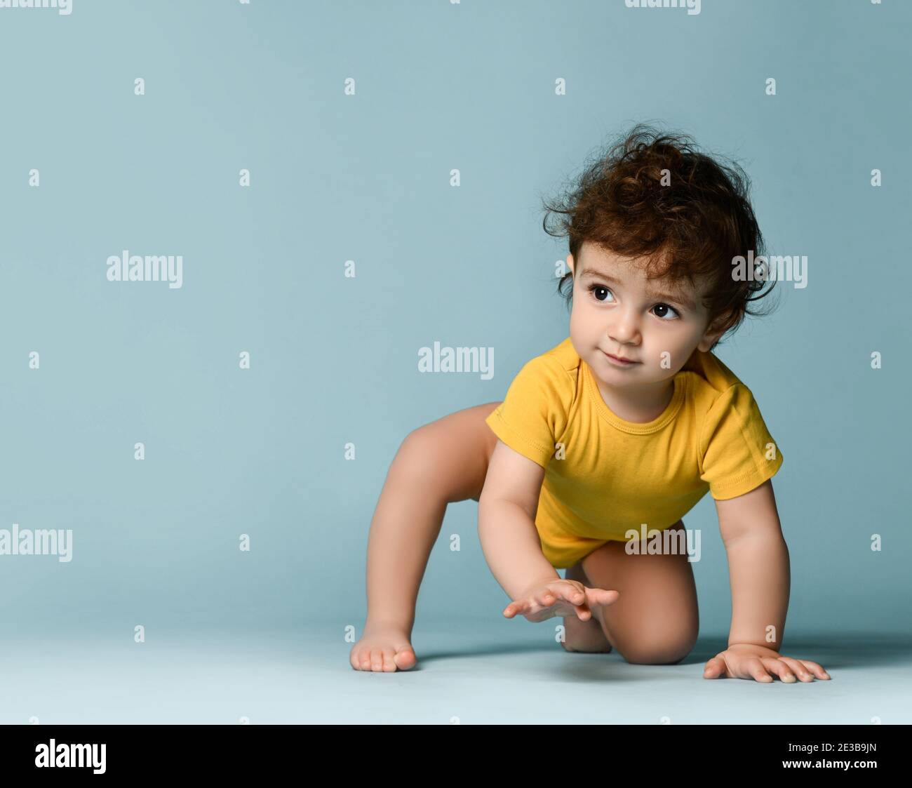 Kleine nette positive lockige Baby junge Kleinkind in gelb bequem Jumpsuit kriecht auf dem Boden und versucht aufzustehen Stockfoto