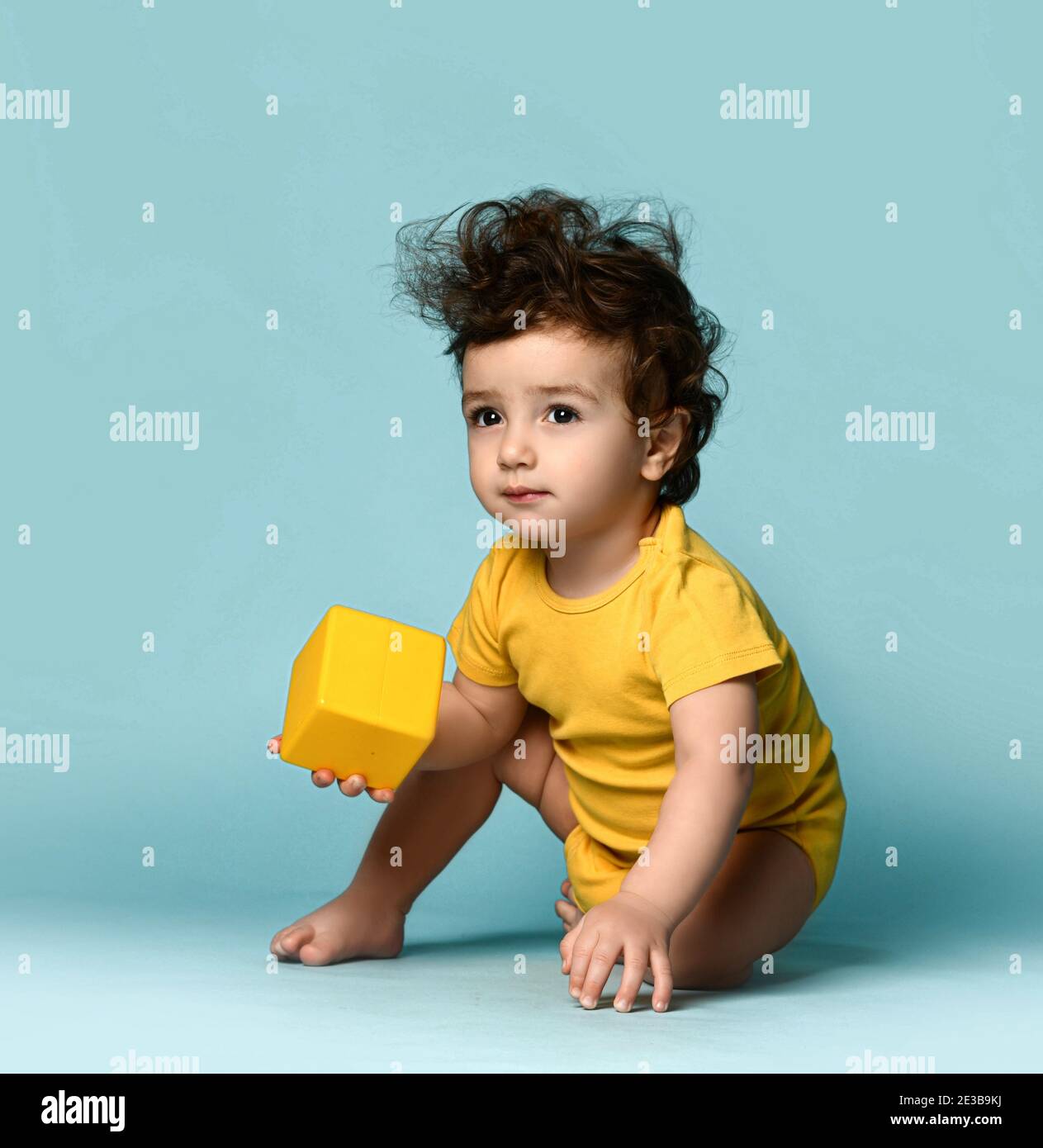 Kleine niedliche lockig haarige Baby junge Kleinkind in gelb bequem Jumpsuit sitzt auf dem Boden und spielt mit gelben Würfel Spielzeug Stockfoto