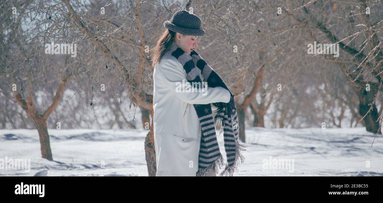 Panoramablick auf eine schöne junge Frau, die warme Kleidung trägt, während sie ihren schwarzen Schal an einem verschneiten Tag legt. Winter Landschaft Hintergrund 2021. Stockfoto