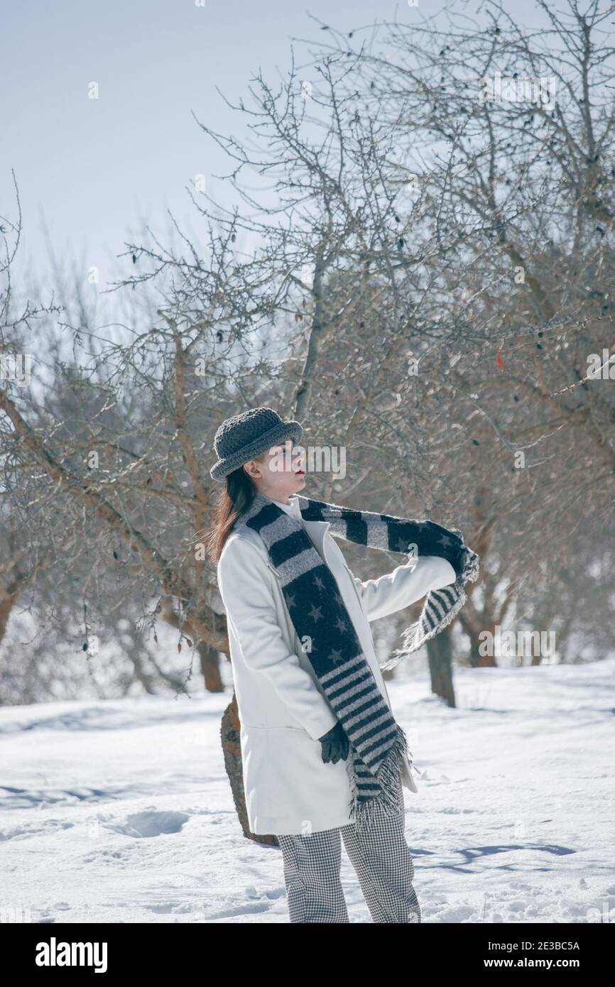 Vertikale Fotografie einer jungen Frau mit Winterhut und Handschuhen, setzt ihren schwarzen Schal an einem verschneiten Tag 2021. Winterlandschaft 2021. Stockfoto