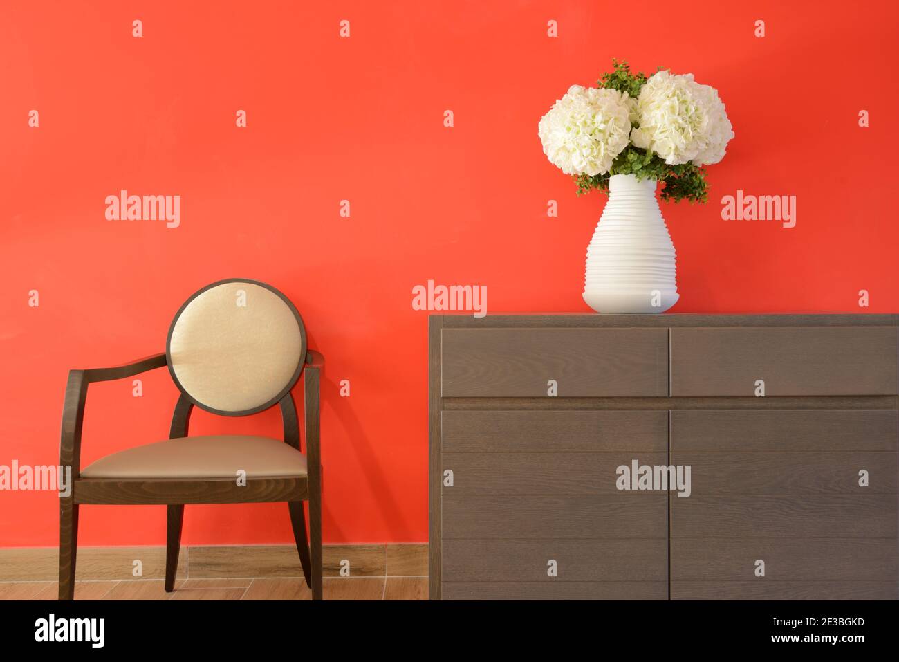 Minimalistisches Interieur mit roter Wand und künstliche weiße Blumen oder Nelken in weißer Vase & moderner Holzstuhl Stockfoto