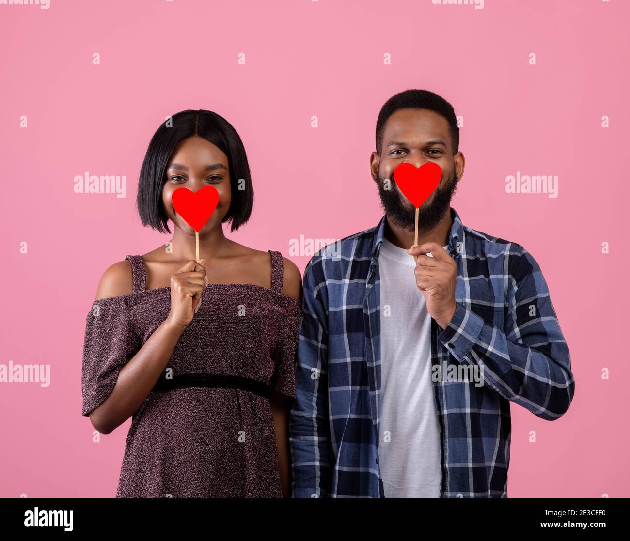 Alles Liebe zum Valentinstag. Leidenschaftliches schwarzes Paar, das ihre Münder mit roten Herzen auf pinkem Studiohintergrund bedeckt Stockfoto