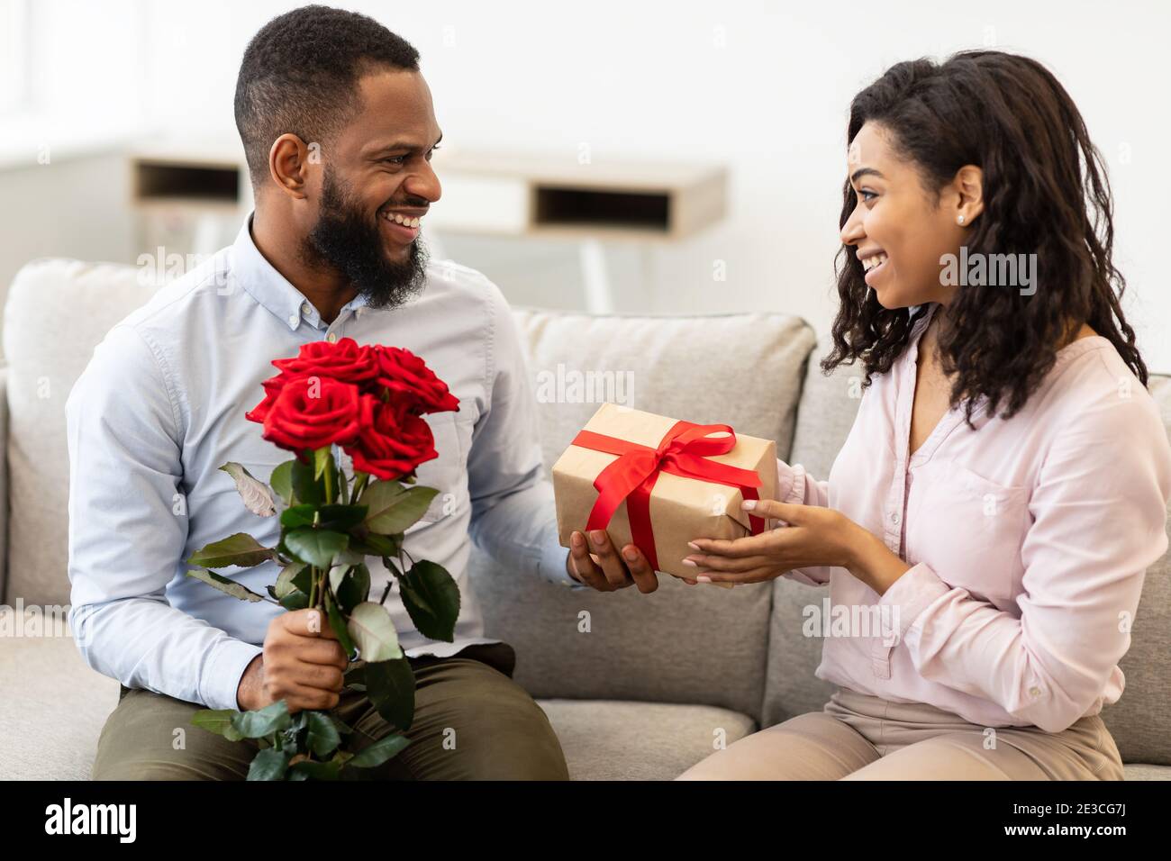 Glücklicher schwarzer Mann, der der Frau rote Rosen und eine Schachtel gibt Stockfoto