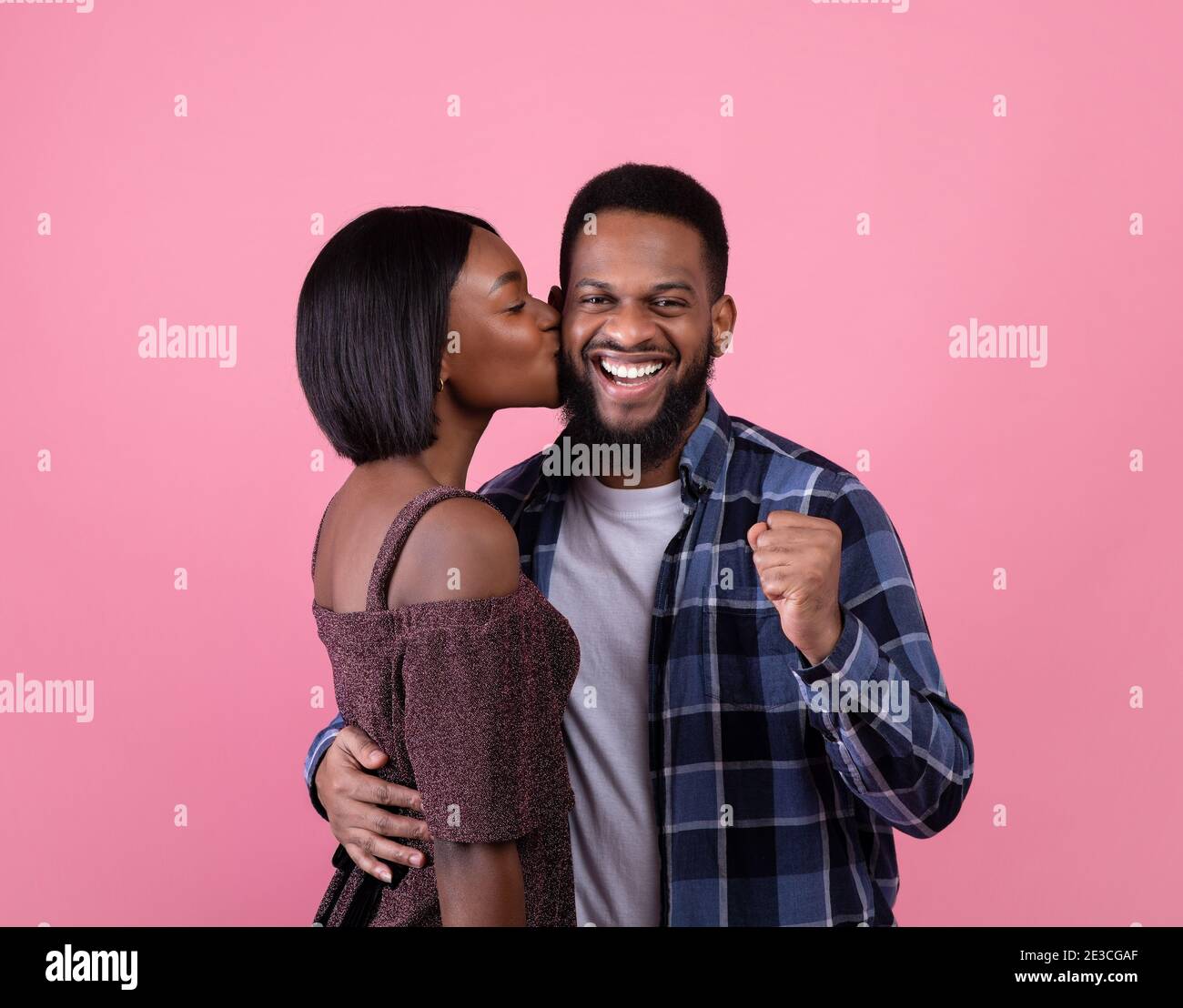 Aufgeregt schwarz Kerl machen JA Geste, junge Freundin küssen seine Wange auf rosa Studio Hintergrund. Valentinstag Stockfoto