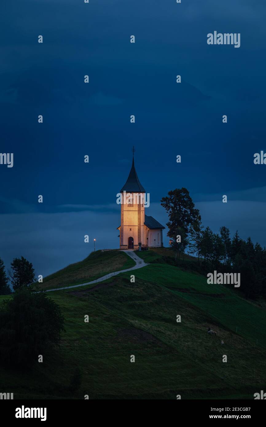 Jamnik, Slowenien - Blaue Stunde in Jamnik mit beleuchteter St. Primoz-Kirche in nebliger Morgendämmerung. Die Julischen Alpen im Hintergrund Stockfoto